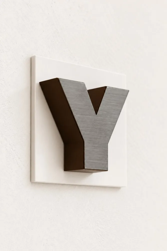 Lettre Y en métal brossé montée sur une plaque blanche fixée au mur