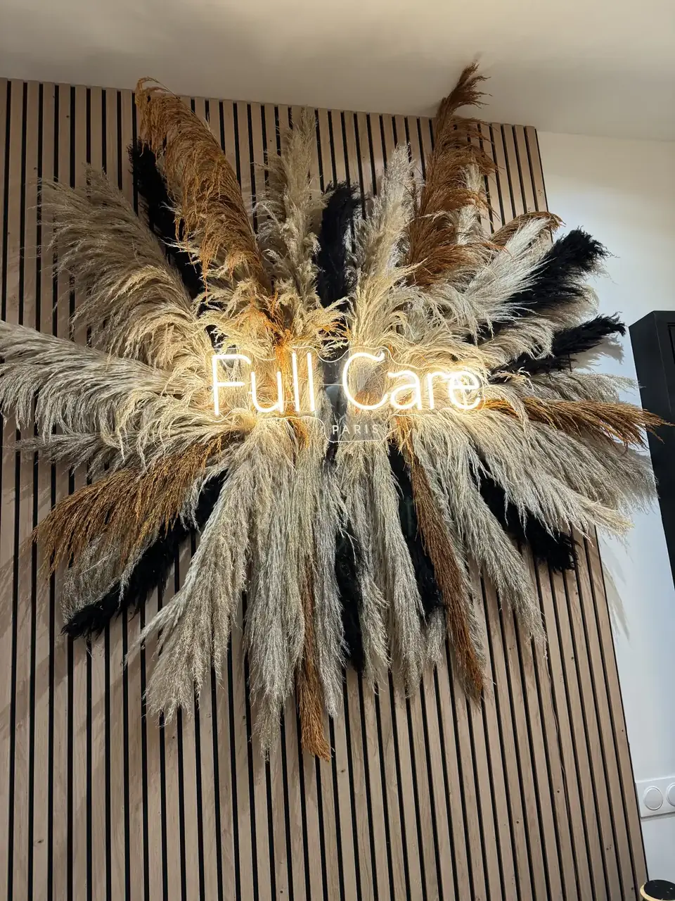 Néon blanc Full Care sur une grande couronne de pampas et plumes sur mur en tasseaux de bois, à Paris 19e, par LADONNA